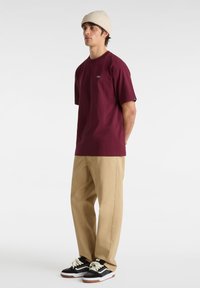 T-shirt en coton bordeaux à manches courtes, avec un petit logo. Associée à un pantalon beige et des baskets noires avec des lacets blancs.