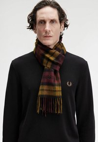 Sciarpa a quadri in ricco bordeaux, giallo e nero, realizzata in morbido tessuto con estremità frangiate. Indossata su un maglione nero a effetto con un emblema.