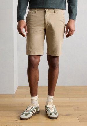 Homme portant un short beige jusqu'aux genoux, des chaussettes blanches et des baskets vertes et blanches, debout sur un sol en bois.