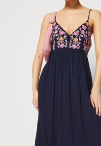 Námořnická modrá maxi šaty s hlubokým výstřihem do V, s barevnou květinovou výšivkou na vrchní části a splývavou sukní. S růžovou kabelkou.