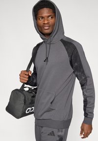 Γκρι φούτερ με κουκούλα και μαύρες διακοσμητικές λεπτομέρειες· το μοντέλο κρατά μαύρη τσάντα duffel με λευκό λογότυπο.