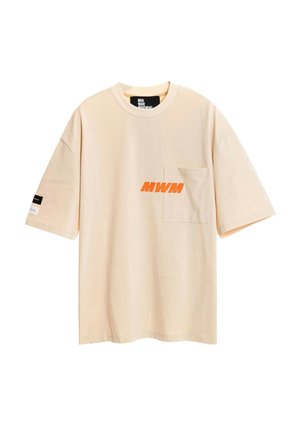 Joven con camiseta oversize beige con logo naranja, pantalones cortos a juego con detalle de cadena, calcetines blancos con texto negro y zapatos beige texturizados.