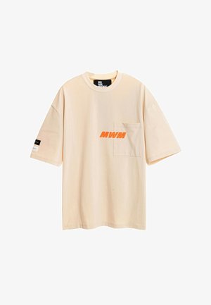 Joven con camiseta oversize beige con logo naranja, pantalones cortos a juego con detalle de cadena, calcetines blancos con texto negro y zapatos beige texturizados.