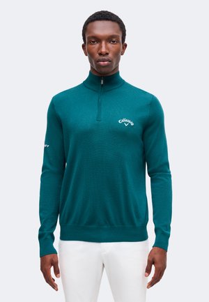 Man met een tealgekleurde trui met een kwartritssluiting met wit Callaway-logo en een witte broek, staand tegen een effen lichte achtergrond.