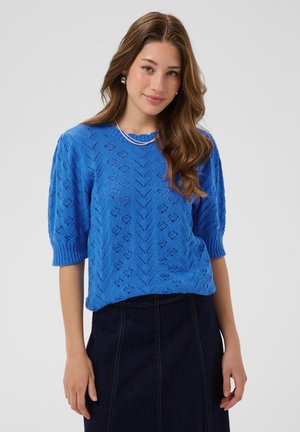 Giovane donna dai lunghi capelli castani indossa un maglione blu brillante a maniche corte, gonna scura, collana d’argento e orecchini a lobo, in piedi all’interno.