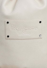 Bolso de piel sintética blanca con una superficie texturizada y un parche de logo de marca en relieve asegurado con remaches de metal plateado.