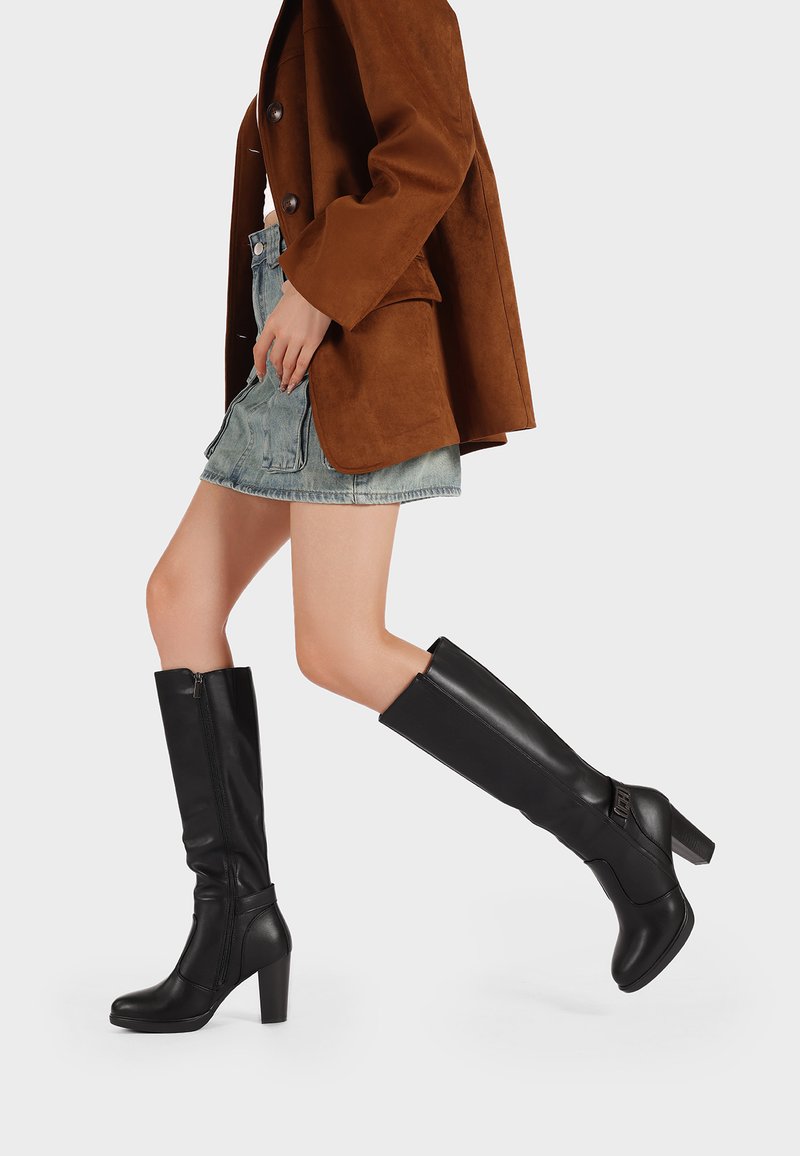 Stivali neri in pelle alta fino al ginocchio con un tacco alto chunky, dotati di una zip laterale e una texture liscia, abbinati a una gonna di jeans e un blazer marrone.