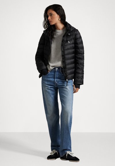 Schwarze gepolsterte Jacke mit Kapuze und Reißverschluss, grauer Pullover darunter, hellblaue Jeans im geraden Schnitt und schwarze Turnschuhe.