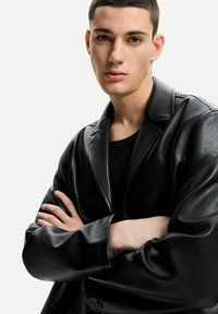 Schwarze Lederblazer mit glatter Textur, breiten Revers und lockerer Passform. getragen über einem schwarzen T-Shirt, mit verschränkten Armen.