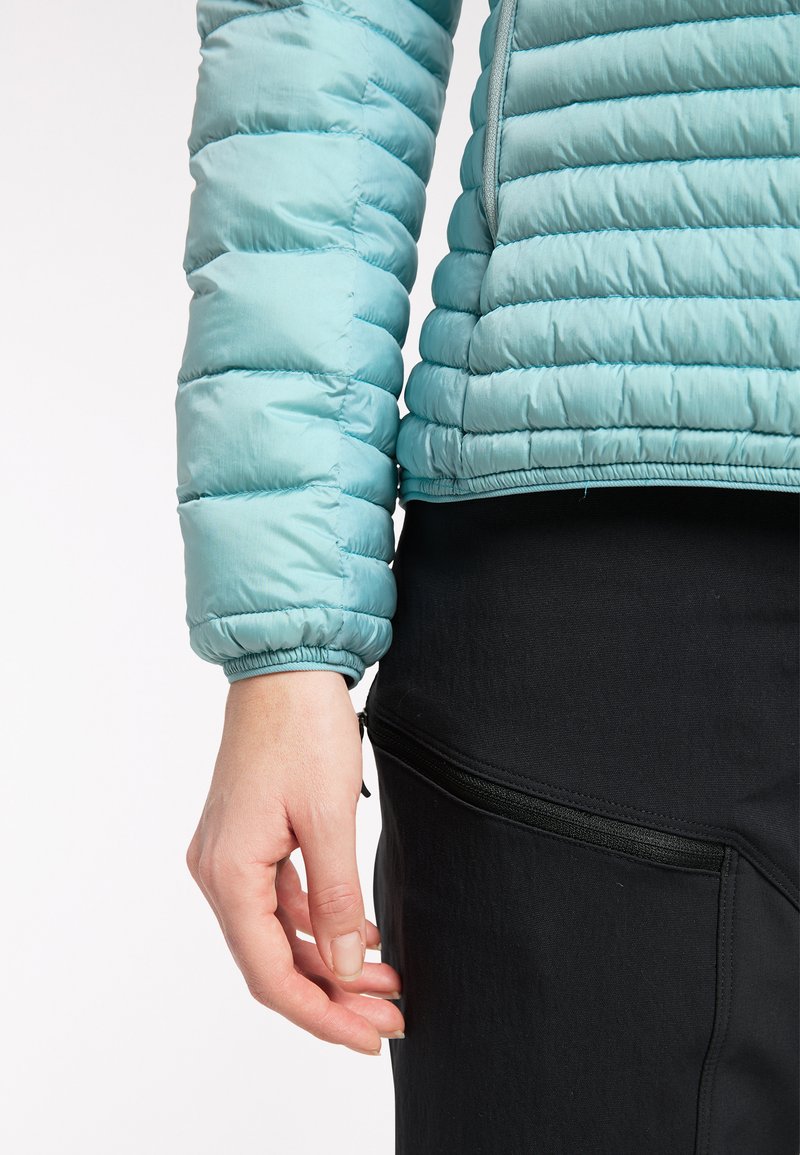 Chaqueta aislante de color azul claro con un diseño acolchado y puños elásticos, combinada con pantalones negros de tela elástica que tienen un bolsillo con cremallera.