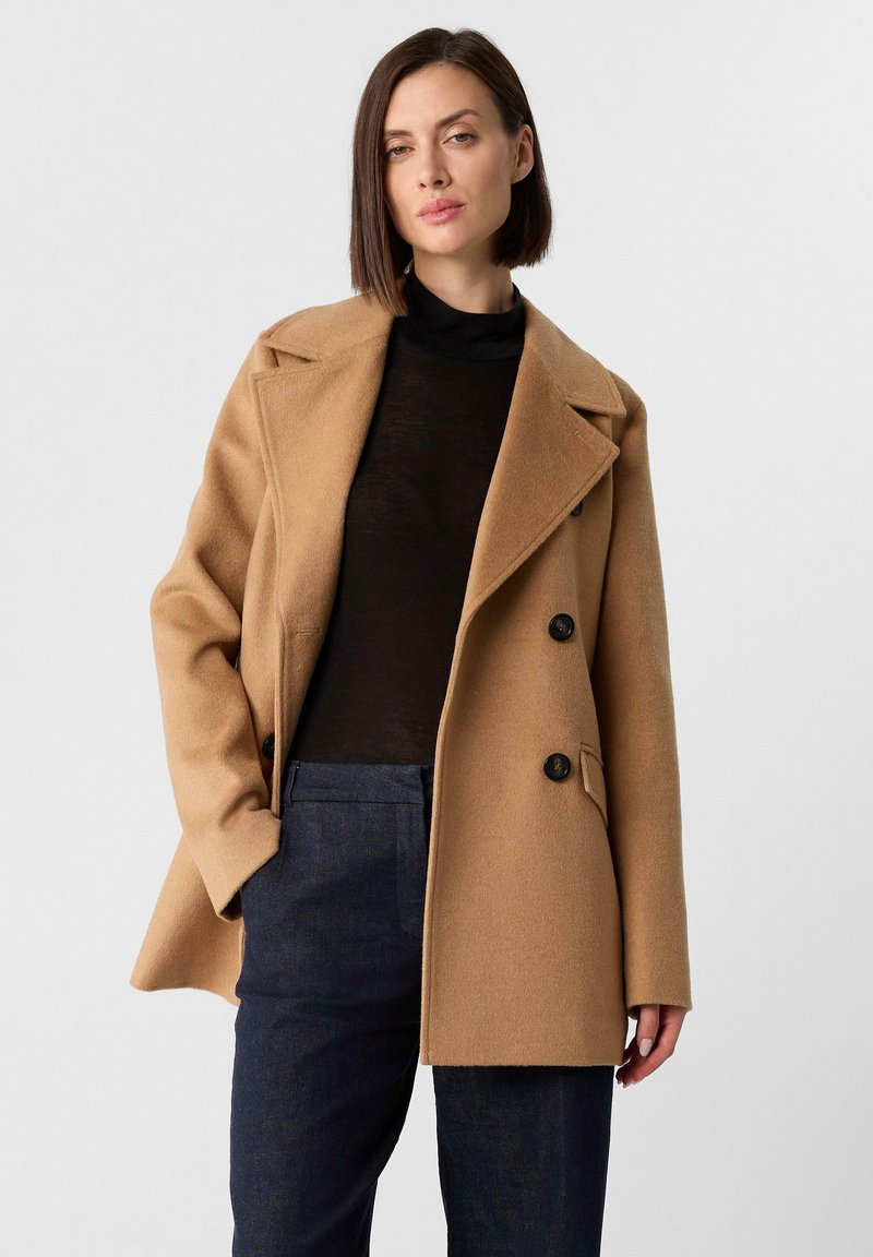 Camel gekleurde double-breasted jas van een zachte wolmix, met notch-lapels, twee voorkruimelpockets en zwarte knopen, gecombineerd met een donkere top en broek.