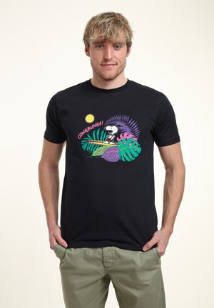 Giovane uomo che indossa una maglietta nera con Snoopy che fa surf e foglie tropicali, con scritta "COWABUNGA!", abbinata a pantaloni verde chiaro.