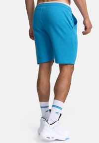 Shorts pour hommes en coton turquoise avec poches latérales, taille élastique et texture lisse ; portés avec des baskets blanches et des chaussettes rayées.