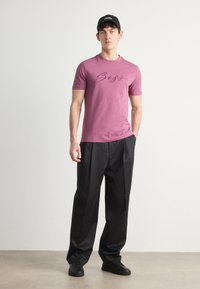 Bordo trumpa rankovė t-shirt su juodu "Boss" logotipu, derinama su juodomis laisvomis kelnėmis ir juodais sportiniais bateliais, visi ant neutralios fono.