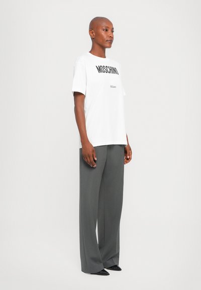 Weißes T-Shirt mit "MOSCHINO" und "Milano" in schwarzer Schrift, kombiniert mit hochtaillierten, weit geschnittenen grauen Hosen und spitzen schwarzen Schuhen.