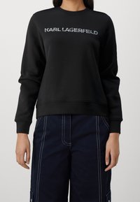 Sudadera negra de manga larga, con puños acanalados y cuello redondo. Presenta un logo en estampado metálico texturizado en el pecho.