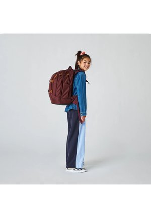 Young girl wearing a large maroon backpack, blue jacket, and two-tone pants, standing and smiling against a plain light background.
