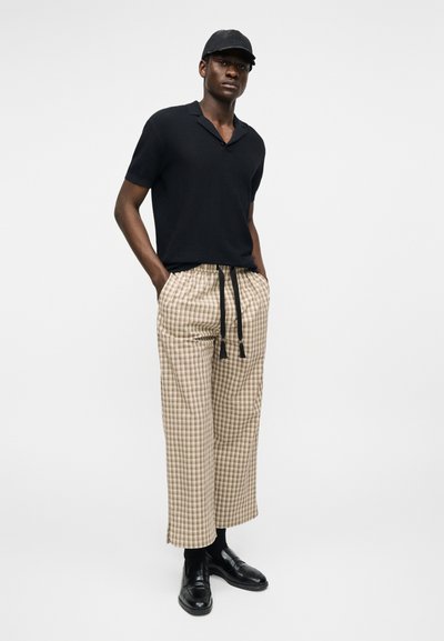 Homme portant une chemise noire à manches courtes, une casquette noire, un pantalon large à carreaux beige avec cordons de serrage noirs, des chaussettes noires et des chaussures en cuir noir, debout.