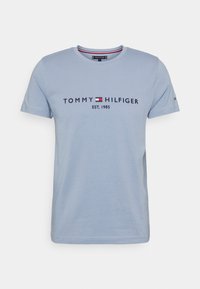 Camiseta de algodón azul claro con mangas cortas, cuello redondo y estampado en el frente que dice "TOMMY HILFIGER EST. 1985" con acentos en azul marino y rojo.