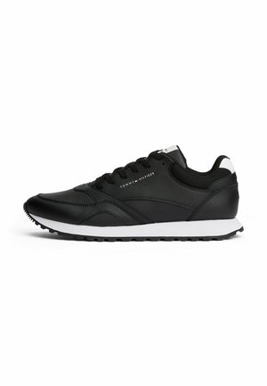 Zwarte synthetische sneaker met een gladde textuur, witte tussenzool, vetersluiting en branding aan de zijkant. Bevat een lichte boog en rubberen zool.