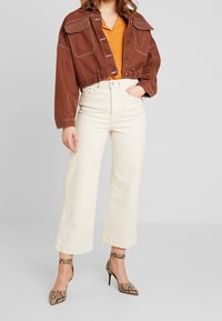 Giacca corta marrone con doppie tasche e dettagli di cucitura, camicia arancione, pantaloni a gamba larga color crema e scarpe col tacco a stampa serpente.