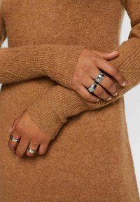 Pull en maille texturée marron avec des manches longues, présentant des poignets côtelés. Plusieurs bagues en argent, dont une avec une pierre verte, aux doigts.