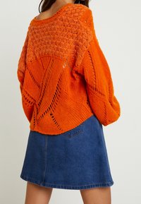 Pomarańczowy sweter na drutach z odkrytym wzorem i teksturowanymi rękawami, zestawiony z ciemną spódnicą jeansową. Sweter ma luźny, krótki krój.