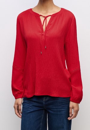 Mujer con blusa roja de manga larga con lazo en el escote delantero y vaqueros azules, de pie frente a un fondo liso.