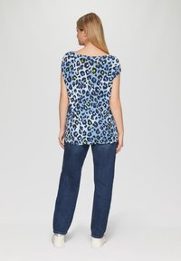 Kurzärmliges, blaues Leopardprint-Top mit Variationen aus schwarzen, gelben und weißen Punkten, kombiniert mit dunklen Jeans und klobigen weißen Schuhen.