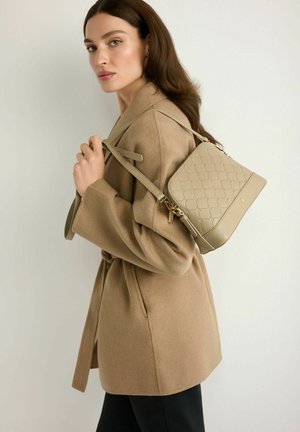 Kobieta w beżowym płaszczu niesie pikowaną beżową torebkę na ramieniu na tle jednolitego jasnego tła.