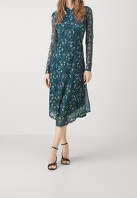 Vestito floreale turchese con maniche lunghe trasparenti, silhouette a linea ad A e orlo all'altezza del ginocchio. Presenta una scollatura incrociata e tacchi neri con cinturini.