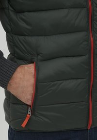 Dunkelgrüne Steppweste mit gepolsterter Textur. Merkmale sind rote Reißverschlussakzente und Taschen. Rippdetails in Grau sind am Arm sichtbar.
