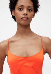 Jasno oranžové saténové šaty s tenkými ramienkami, hlbokým výstrihom a priliehavým živôtikom. Hladká textúra s geometrickým švovým detailom.