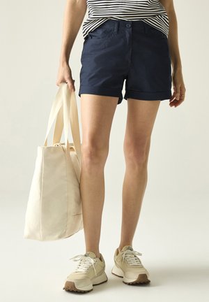 Personne portant un short bleu marine et un haut rayé tenant un grand sac fourre-tout crème, debout en baskets beiges sur fond uni.