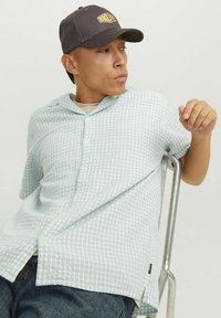 Chemise à manches courtes, à carreaux vert menthe avec col, dotée d'une fermeture à boutons et d'une fente sur le côté. Portée avec une casquette sombre, assise sur une chaise en métal.
