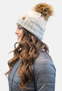 Bonnet tricoté gris clair avec un motif texturé, doté d'un pompon en fausse fourrure sur le dessus et d'un patch en cuir sur le côté.