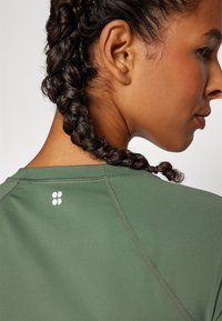 Zelené sportovní tričko s hladkým, texturaovaným materiálem. Má kulatý výstřih, raglánové rukávy a malé bílé logo na zadní straně.