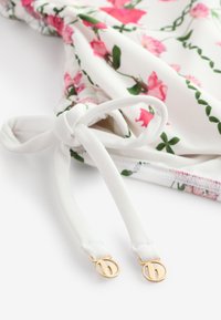 Tessuto bianco con stampa floreale rosa legato a un nodo, mostrando spalline bianche che terminano con anelli di metallo dorati con dettagli della lettera B.