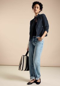 Marineblazer, schwarzes Top, hellblaue Jeans und schwarze flache Schuhe. Trägt eine schwarz-weiß gestreifte Tragetasche. Neutraler Hintergrund.