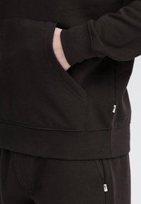 Sweatshirt marron foncé avec une poche kangourou et des poignets côtelés. Présente une étiquette latérale et un tissu doux et texturé. Détails de couture clairs.