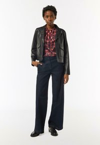 Czarna skórzana kurtka, bluzka w czerwono-czarny wzór, szerokie ciemnoniebieskie dżinsy, czarne buty na gumowej podeszwie, ręce w kieszeniach.