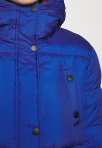 Blue padded jacket with a high collar, featuring black snap buttons and a side pocket. Smooth, shiny texture with subtle wrinkle details.