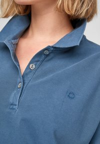 Modrá bavlněná polo košile s texturovaným povrchem, dvouknoflíkovou légou a malým logem na hrudi. Má uvolněný límec.