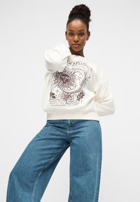 Φούτερ κρεμ με μεγάλο καφέ γραφικό σχέδιο ενός ήλιου και φεγγαριού, συνδυασμένο με ανοιχτό μπλε τζιν παντελόνι φαρδιάς γραμμής.