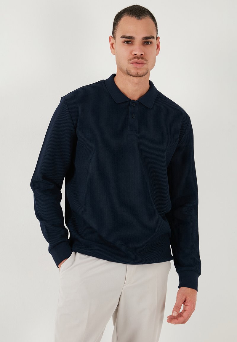 Tricou polo cu mânecă lungă de culoare bleumarin, cu un design texturat în dungi, cu închidere cu trei nasturi și guler tradițional. Purtați-l cu pantaloni de culoare deschisă.