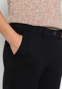Pantalon noir sur mesure avec poches latérales, texture lisse, et fermeture à boutons à la taille. Porté avec un haut en maille multicolore.