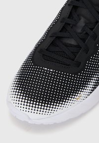 Shoe atlético preto com parte superior em malha apresentando um padrão de bolinhas em branco, e uma sola branca. Atacadores e detalhes em preto. Logotipo em dourado.
