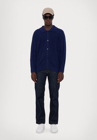 Navy knitted cardigan with a collared neckline and front buttons, paired with dark jeans and white sneakers. Accessorized with a beige cap and sunglasses.