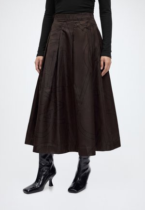 Femme portant une jupe midi trapèze marron foncé avec de grandes lettres subtiles et des bottines en cuir verni noir à talons.
