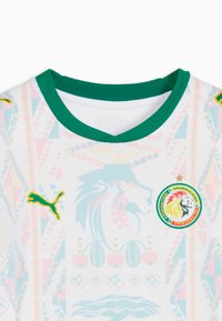 Maillot de sport blanc avec col vert, logos Puma jaunes, écusson de la fédération de football du Sénégal, et motifs géométriques colorés subtils ainsi que des motifs de lion.